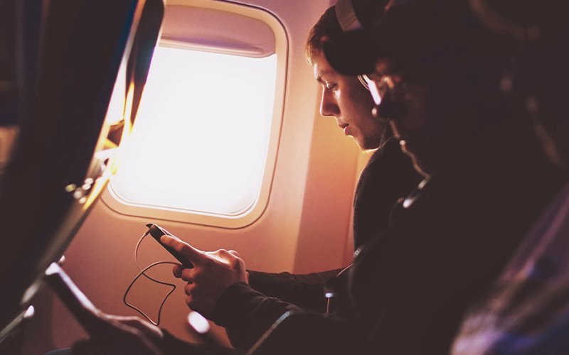 An image shows people sitting in the airplane. One of the persons is sitting by the window while looking at the cell phone.