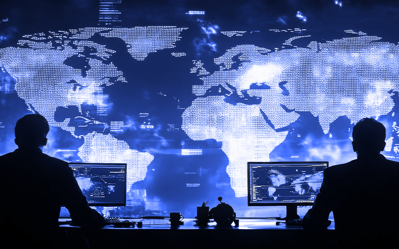 Two silhouetted individuals work at computer stations in front of a large digital world map with glowing blue data points and network connections.