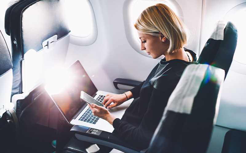 An image of a woman sitting in an airplane, working on her laptop while looking at the screen of her phone.