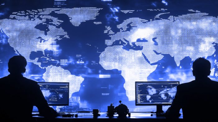 Two silhouetted individuals work at computer stations in front of a large digital world map with glowing blue data points and network connections.