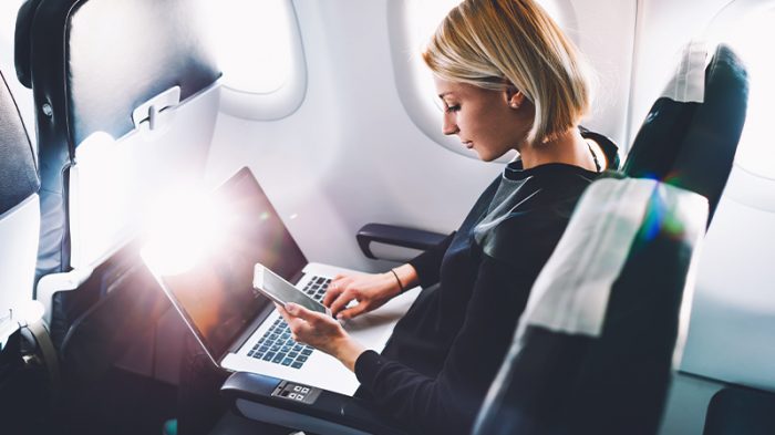 An image of a woman sitting in an airplane, working on her laptop while looking at the screen of her phone.