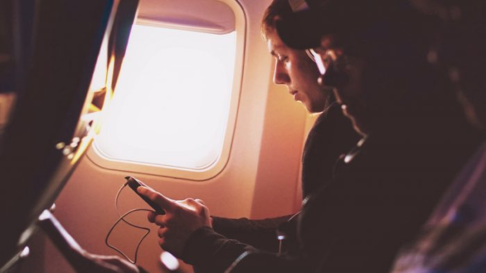 An image shows people sitting in the airplane. One of the persons is sitting by the window while looking at the cell phone.